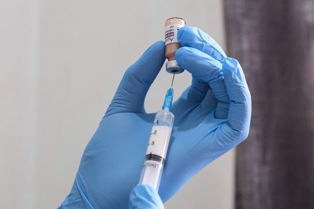 A pair of hands wearing blue, medical gloves hold a vial of liquid and a syringe. They are extracting liquid from the vial into the syringe.