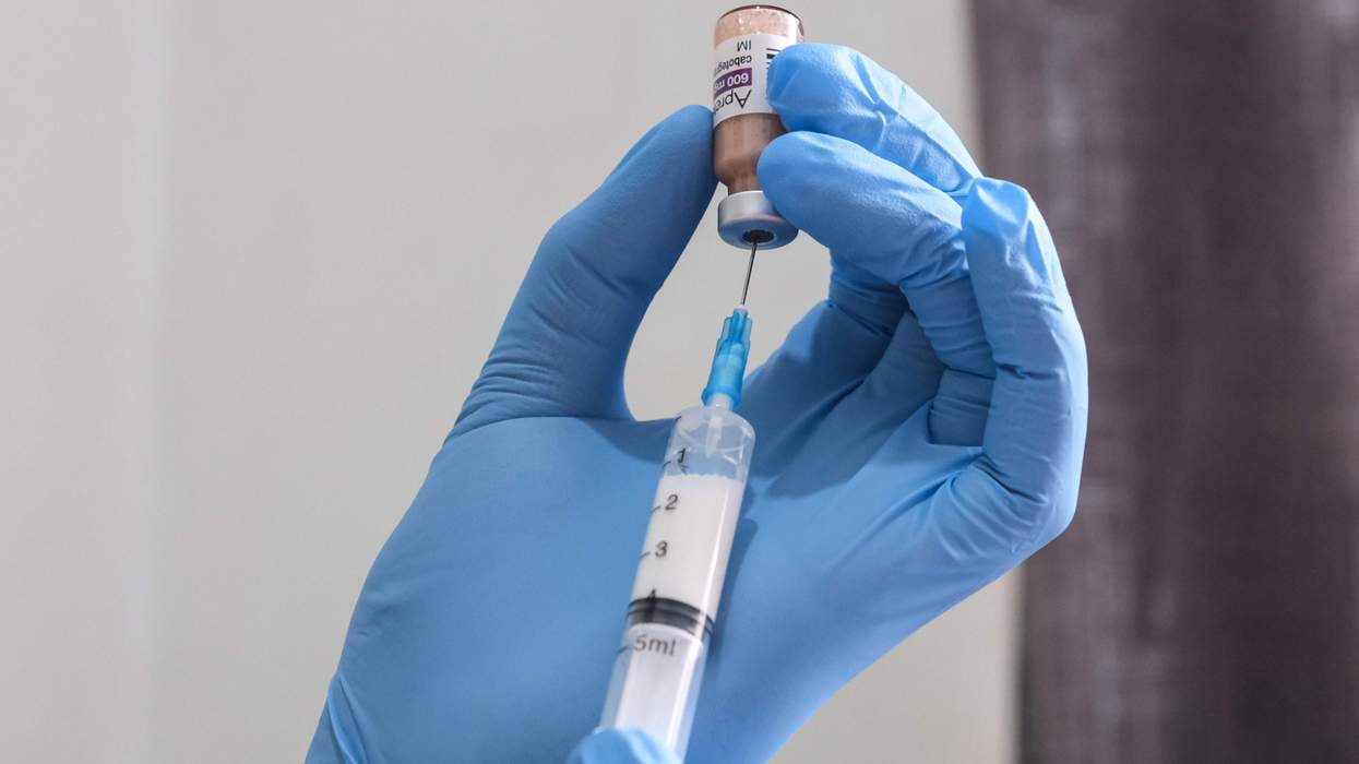 A pair of hands wearing blue, medical gloves hold a vial of liquid and a syringe. They are extracting liquid from the vial into the syringe.