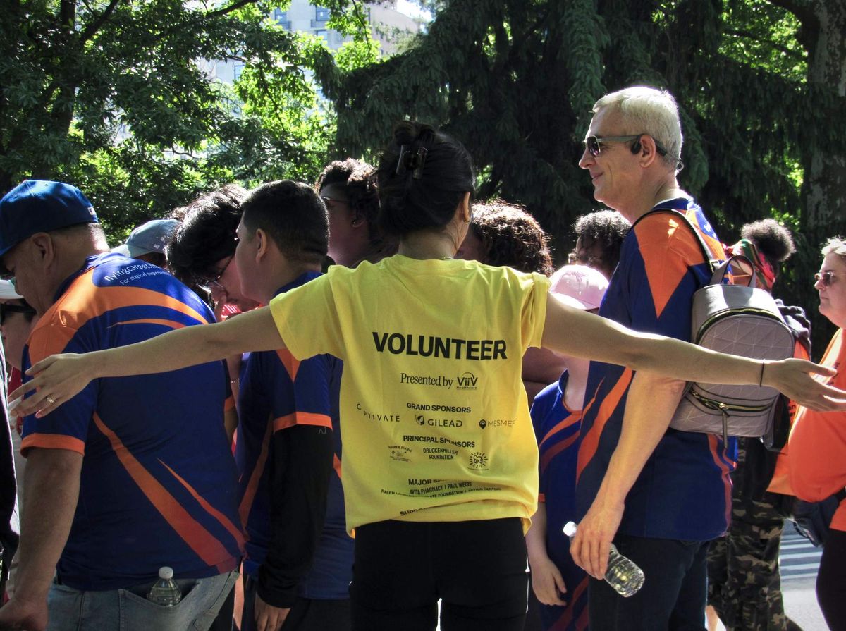A person in a yellow "volunteer" shirt waves a crowd of walkers through a park.